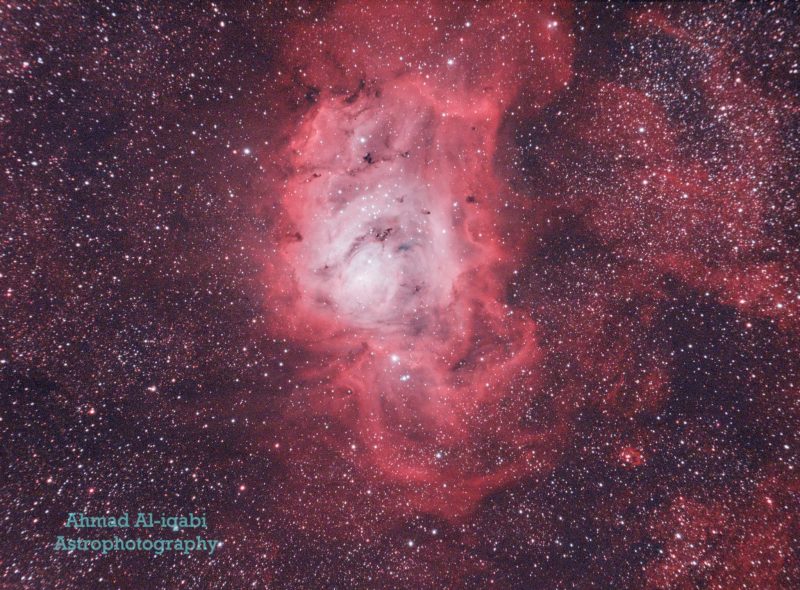 Large swirl of reddish and light blue nebulosity in a dense star field.