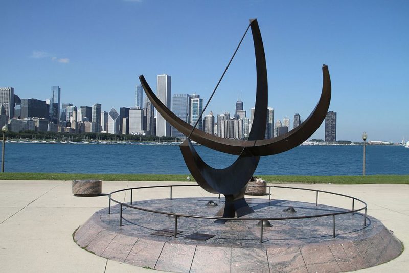 Longest days: Giant sundial made of 2 intersecting semicircular bars, with city skyline beyond.
