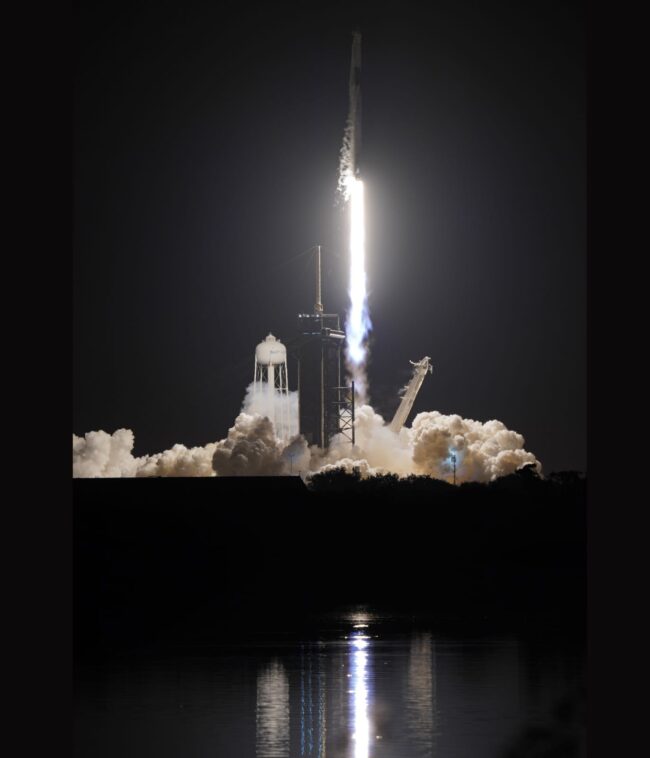 Rocket launch reflected in water. There is  grey smoke at the bottom of the rocket.