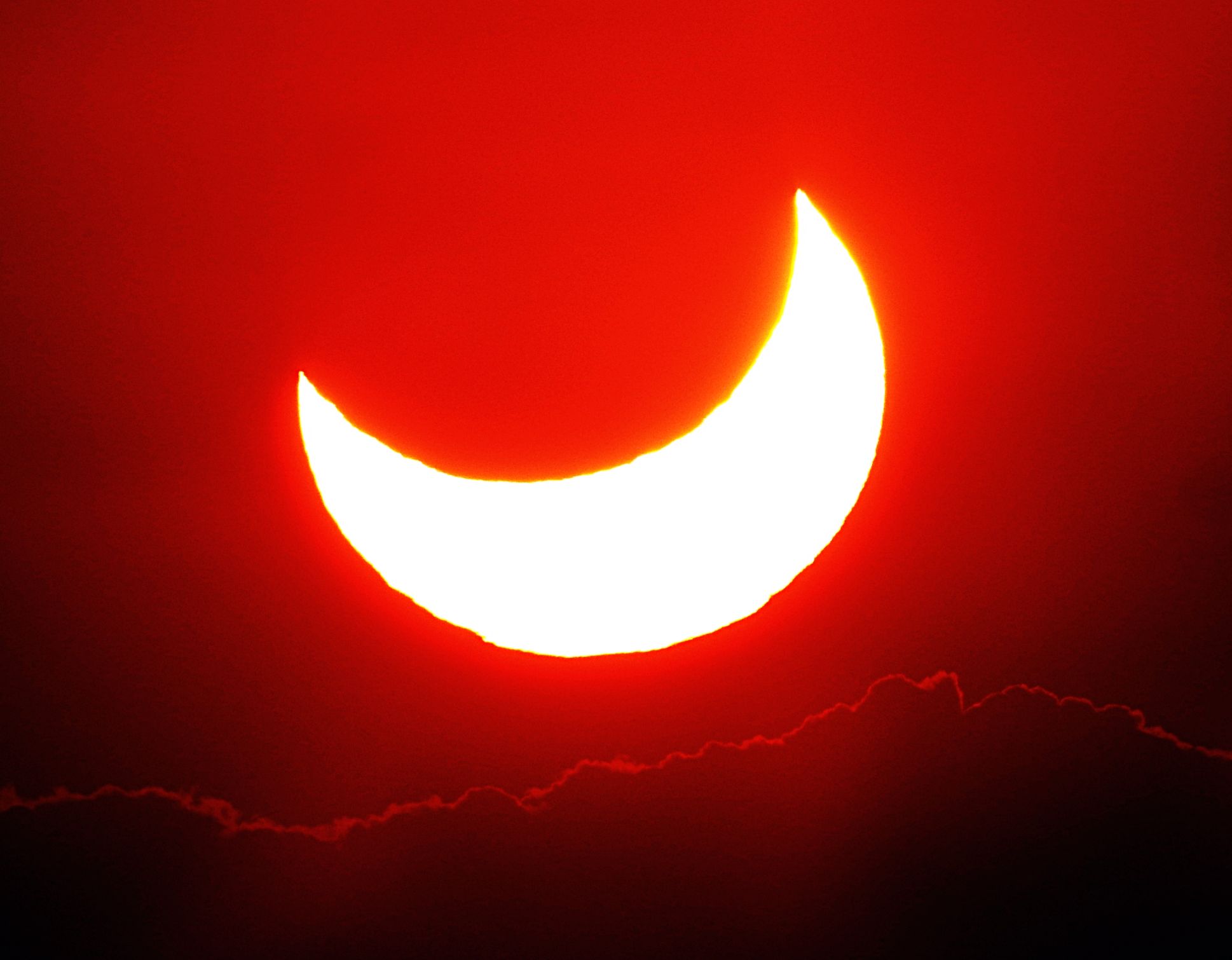 A partially eclipsed sun, above clouds. There are some clouds below and a red haze.