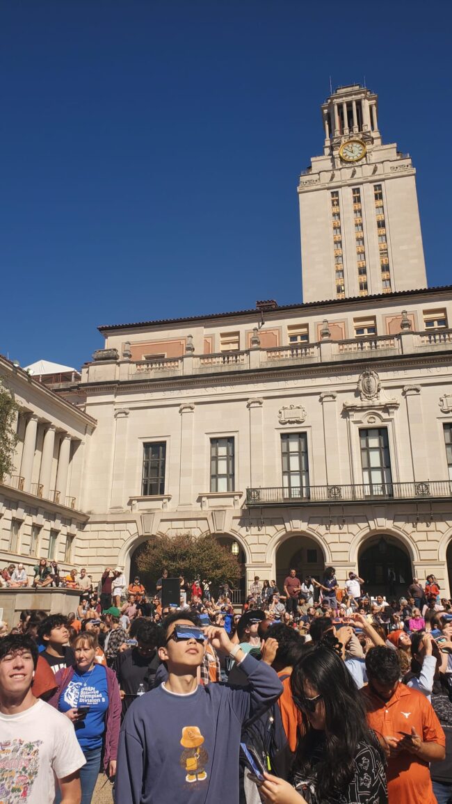 A crowd of people on a college campus, many wearing eclipse glasses.