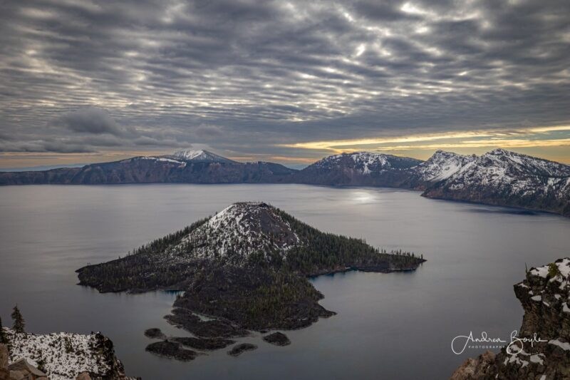 A chain of mountains around a water surface. There is another mountain in the middle of the immage, surrounded by water. There is a grey sky covered in clouds. There are some bright areas in the sky, one of them reflects in the water, creating a crescent shape.