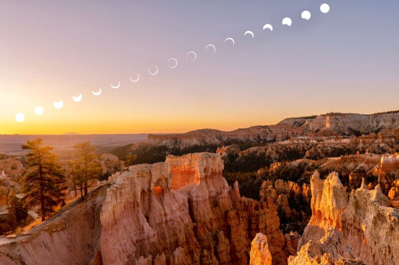 Landscape with mountains and peaks and some trees at the left side. There are 17 different shaped for the eclipsed sun from bottom left to top right.