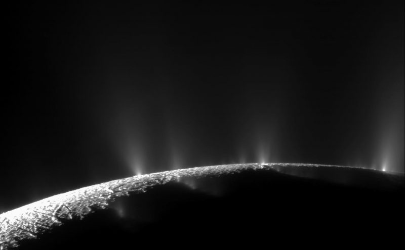 Dark, gray surface of a moon with bright jets pointing upwards.