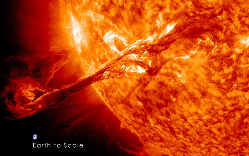 Carrington Event: Section of orange-yellow mottled sun, with long, looped glowing streamer coming out. Tiny Earth beside it.