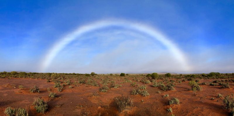 Nearly semicircular fuzzy white arc with very faint colors over a red-soil desert landscape.