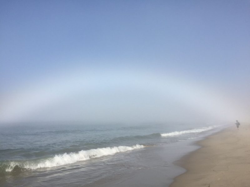 Beach with low foamy waves rolling in under a fuzzy white arc, a fogbow.