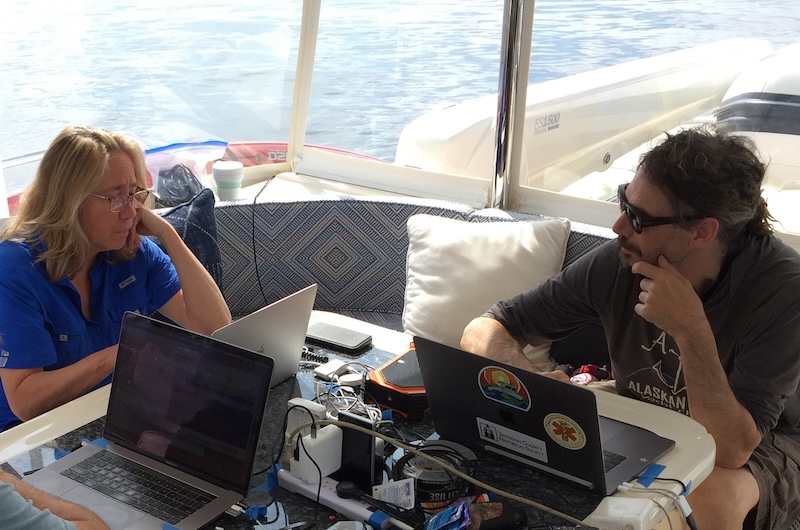 Woman and man on a boat shown at a table using laptops.