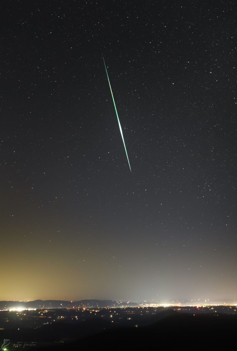Long, bright, green streak in a starry sky over a city.