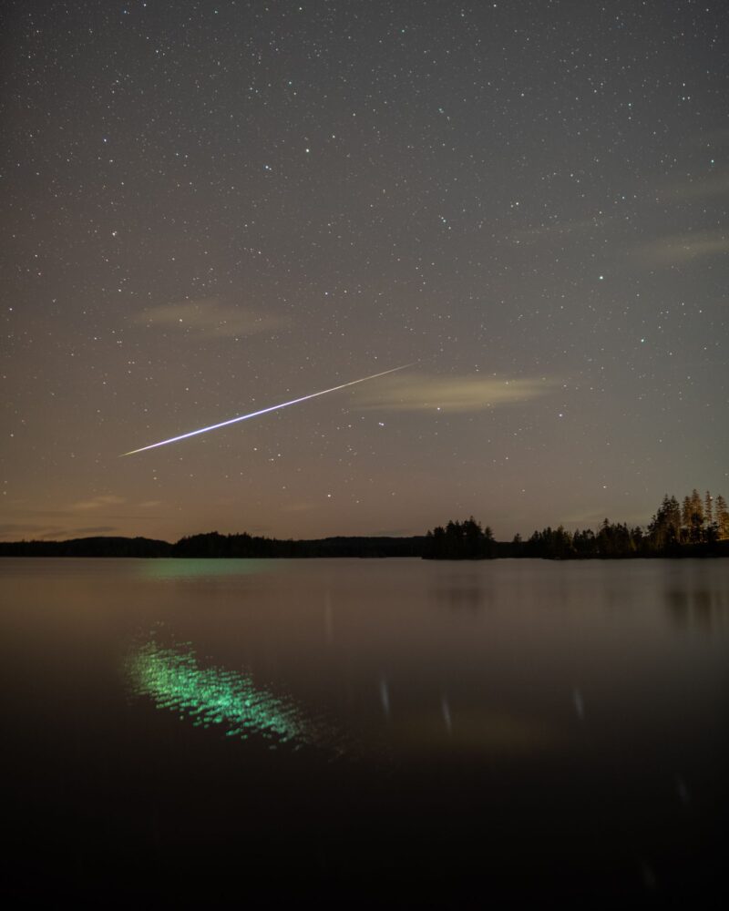 Geminid: Starry sky with a white, long streak crossing it. It is reflected in the water in a green color.