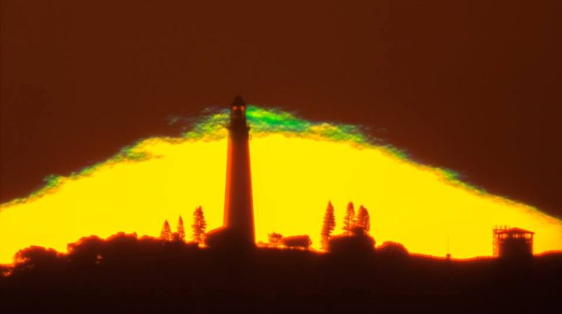 Silhouette of tower and trees against wide, yellow setting sun mostly under horizon with green streak at top.