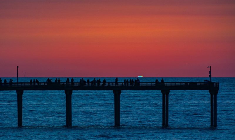 Green bit of light at horizon above dark water with an orange sky and a pier in the foreground.