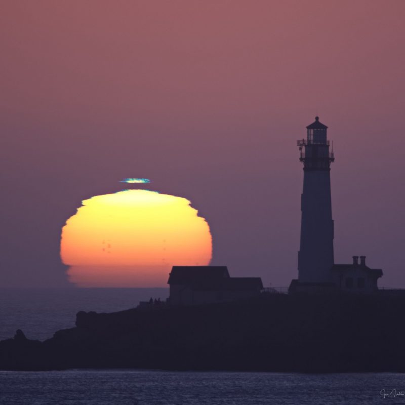 Sun on the horizon with a short blue streak above it and a silhouetted lighthouse on the right.