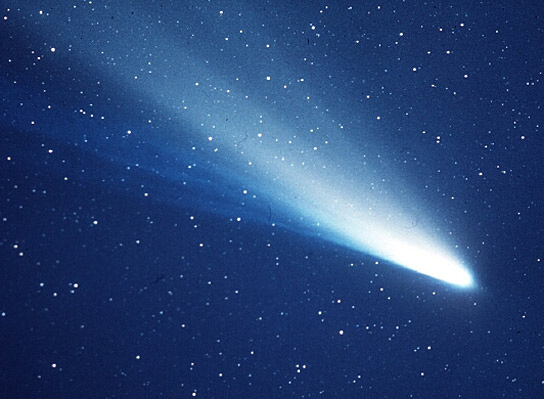 Bright white comet with wide glowing tail streaming out from it in starry deep blue sky.