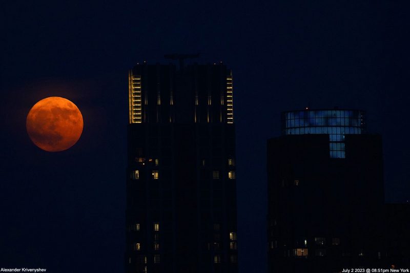 Tops of 2 tall buildings at night with deep orange moon behind.