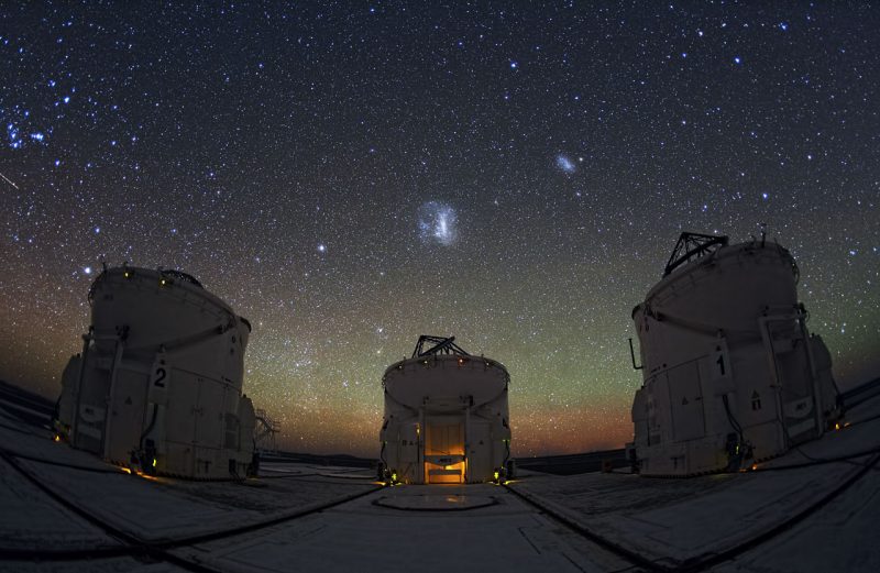 Three dish telescopes under dark starry sky with large puffy white spot at center of sky and smaller one to the upper left.