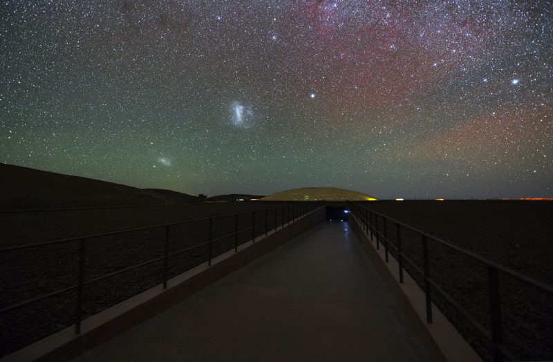 Very starry sky with a large fuzzy patch and a smaller fuzzy patch to lower left above a road.