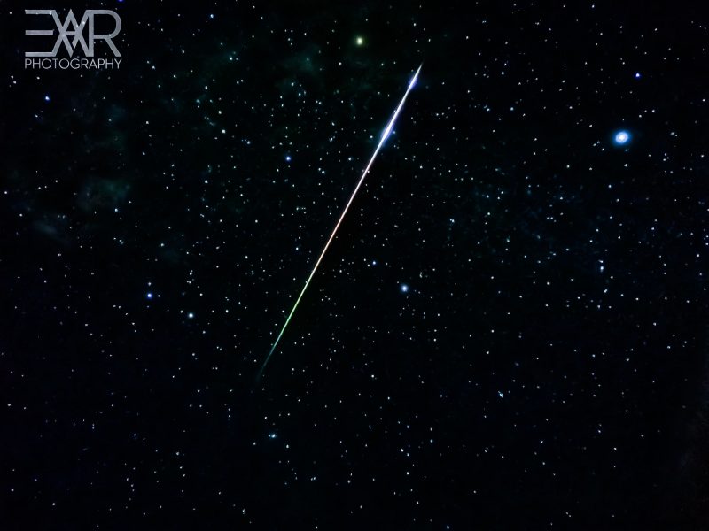 Long, bright, colorful (green, red and blue) meteor in starry black sky.