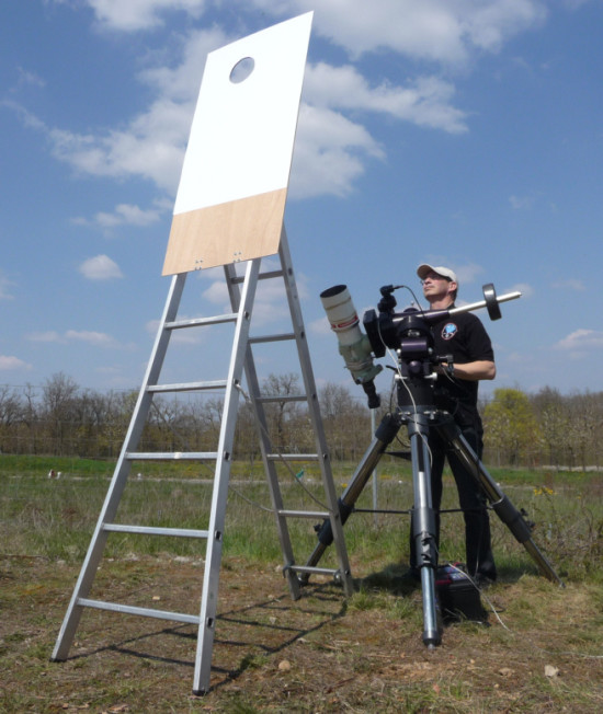 Man standing with telescope looking at sunshade with a hole for viewing the moon in the daytime sky.