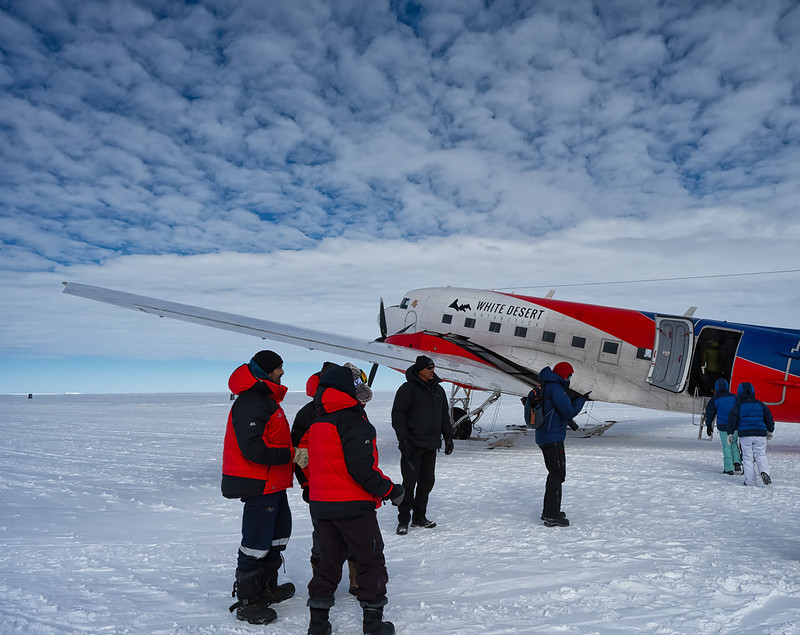 6 persons with big coats in front of a red, blue and whote plane. The ground is covered in snow.