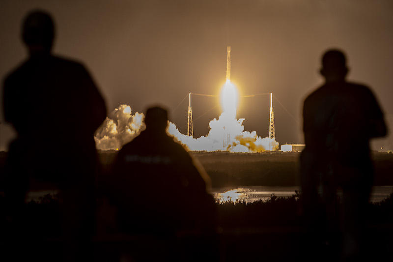 Starlink: A narrow rocket, atop brilliant white flames, launches into a golden night sky. 3 people silhouetted in foreground.