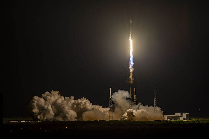 Starlink: A narrow rocket, atop golden flames, launches upward into a black night sky while white clouds billow below.