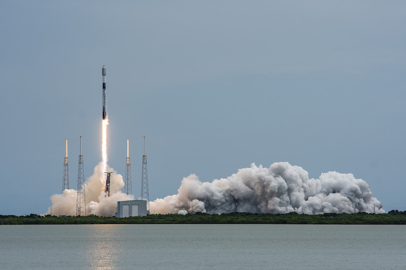 Starlink: Across a body of water, a white and black rocket launches into a blue sky, above billowing clouds of smoke.