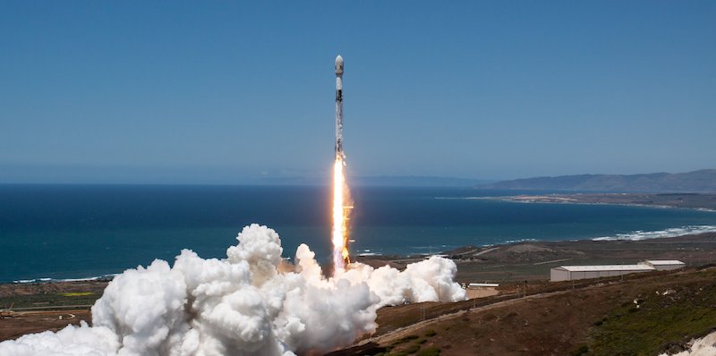 Starlink: A white and black cylindrical vehicle launches upward into a blue sky, leaving behind clouds of smoke on the brown ground.