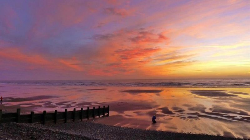 Orange sunset over beach with waves coming in & long wooden structure sticking out into the sea.