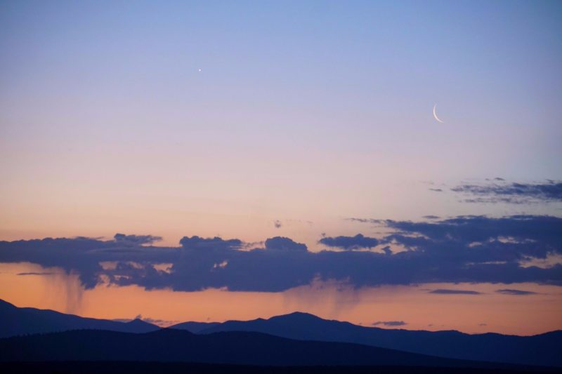 Thin crescent moon, Venus, virga coming from a single stripe of cloud against dawn sky, over dark mountains.